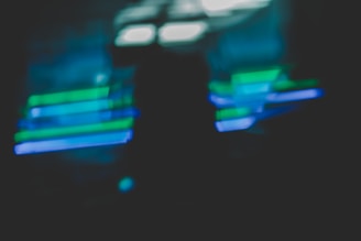 A blurred image featuring overlapping horizontal streaks of neon green and blue light against a dark background, creating an abstract and dynamic visual effect.