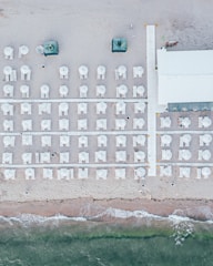An aerial view of a beach with neatly arranged rows of white beach umbrellas and lounge chairs on the sand. A wooden walkway divides the area, leading to a building on the right side. The shoreline with gentle waves meets the sand at the bottom of the image.