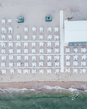 An aerial view of a beach with neatly arranged rows of white beach umbrellas and lounge chairs on the sand. A wooden walkway divides the area, leading to a building on the right side. The shoreline with gentle waves meets the sand at the bottom of the image.
