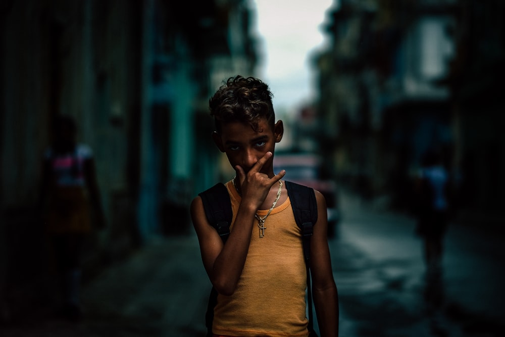 boy in orange tank top and black backpack standing in alley