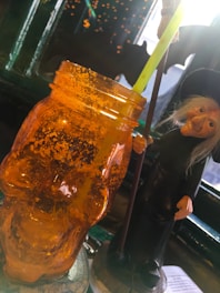 An orange skull-shaped jar with a straw protruding from it, positioned beside a figure of a witch holding a broomstick. In the background, there are lights and a dark silhouette of a bat. The setting gives off a Halloween-themed ambiance.