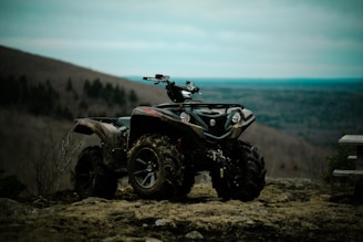 An all-terrain vehicle is parked on uneven, rocky terrain. The vehicle is heavily built, designed for off-road use, and its tires are covered in mud. In the background, there is a vast landscape of rolling hills and a cloudy sky, suggesting a remote, natural setting.