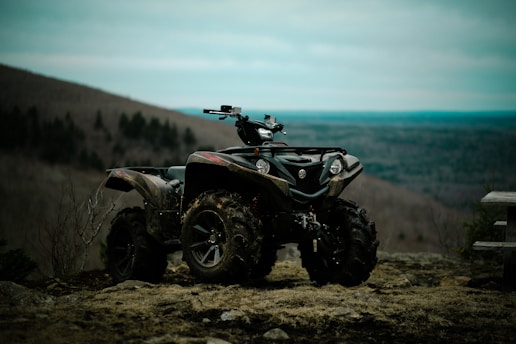 An all-terrain vehicle is parked on uneven, rocky terrain. The vehicle is heavily built, designed for off-road use, and its tires are covered in mud. In the background, there is a vast landscape of rolling hills and a cloudy sky, suggesting a remote, natural setting.