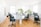 A bright and spacious office environment with several people working at desks. The room features large windows allowing natural light to flood in, wooden floors, and modern office chairs. Two large green plants are placed on the central desk, adding a touch of nature to the space.