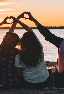 four people sitting on shore forming hearts with their hands during golden hour