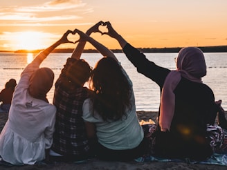 four people sitting on shore forming hearts with their hands during golden hour