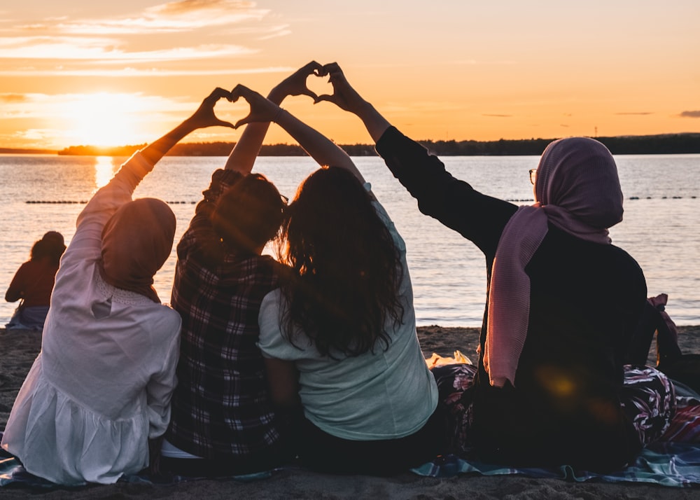 four people sitting on shore forming hearts with their hands during golden hour