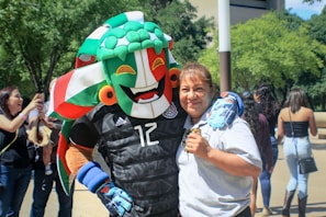 A person joyfully embraces a costumed mascot character with a vibrant headpiece in a public setting. The background features other people, some of whom are taking photos, and trees with a building visible.