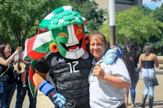 A person joyfully embraces a costumed mascot character with a vibrant headpiece in a public setting. The background features other people, some of whom are taking photos, and trees with a building visible.