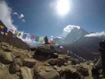 A mountainous landscape with a clear blue sky and bright sunlight overhead. Colorful prayer flags stretch across the scene, fluttering in the wind. Majestic snowy peaks tower in the background, while rocky terrain makes up the foreground. A person is present, possibly taking a photo, beside some rocks.
