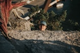 A young person wearing a cap peeks over a rocky ledge. Behind them, a dry, twisted tree branch and a blurred background with distant landscape and water create a rugged outdoor setting.