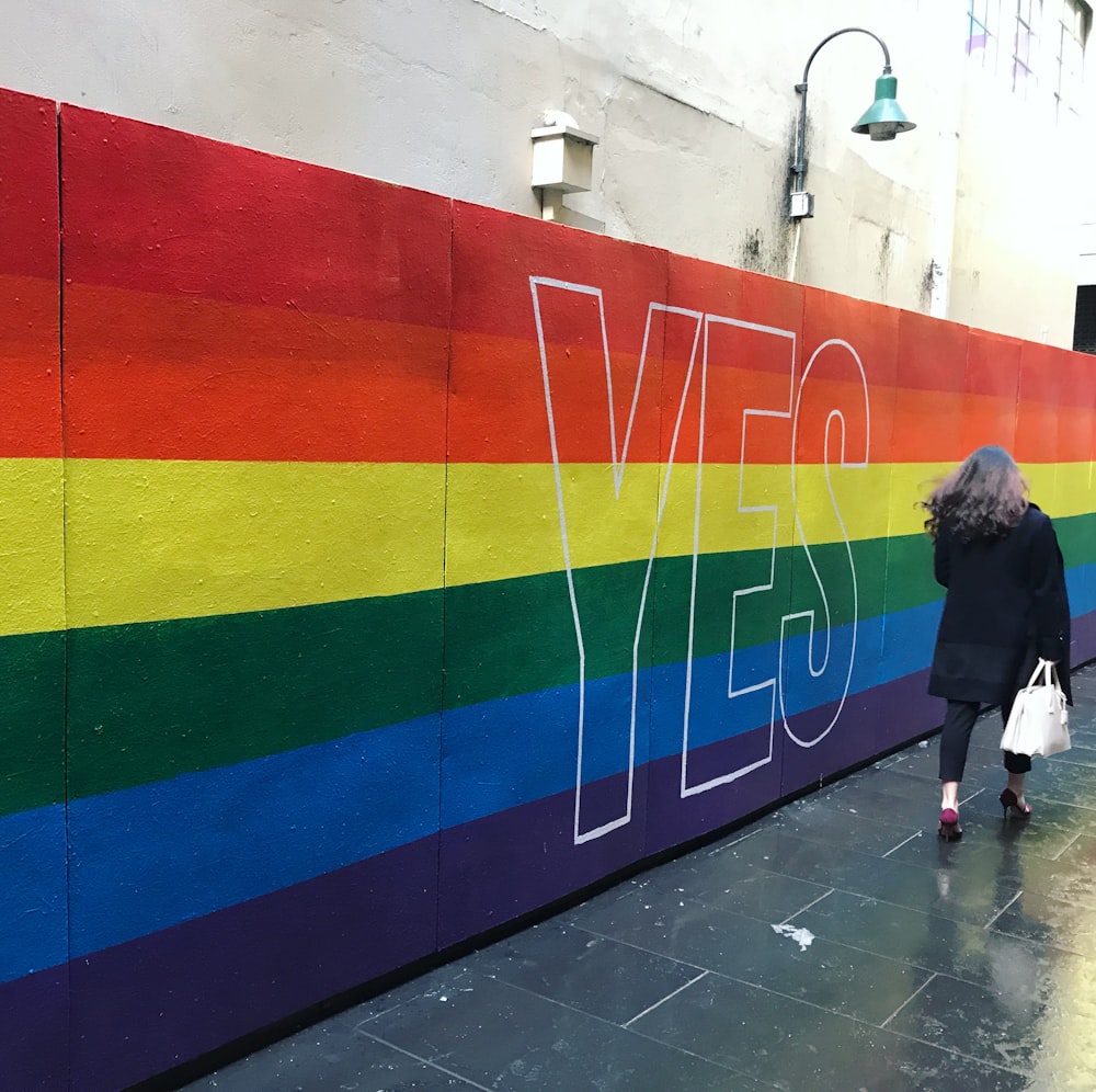 Una mujer caminando por una acera más allá de una pared pintada de arco iris