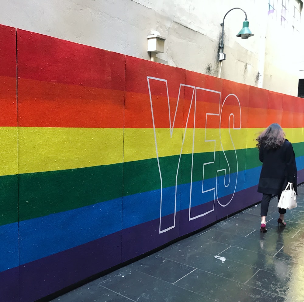 a woman walking down a sidewalk past a rainbow painted wall