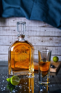A bottle of tequila labeled 'Seleccion Suprema Extra Añejo' is placed on a reflective surface with scattered salt crystals. Beside it, there's a glass containing some of the spirit. A sliced lime and a lime wedge rest nearby on a wooden cutting board, while a dark blue cloth drapes in the background against a wooden wall.