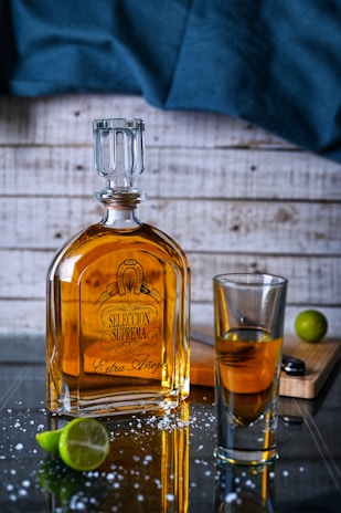 A bottle of tequila labeled 'Seleccion Suprema Extra Añejo' is placed on a reflective surface with scattered salt crystals. Beside it, there's a glass containing some of the spirit. A sliced lime and a lime wedge rest nearby on a wooden cutting board, while a dark blue cloth drapes in the background against a wooden wall.