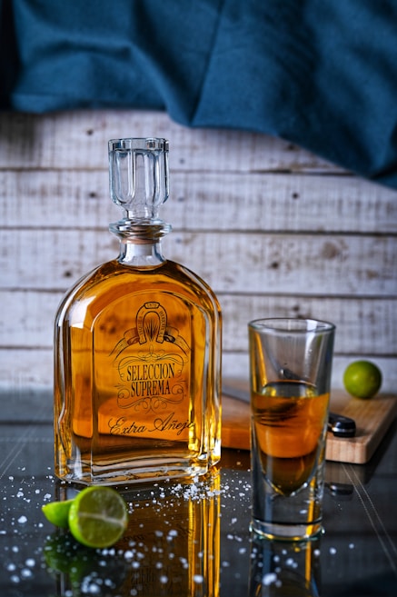 A bottle of tequila labeled 'Seleccion Suprema Extra A&ntilde;ejo' is placed on a reflective surface with scattered salt crystals. Beside it, there's a glass containing some of the spirit. A sliced lime and a lime wedge rest nearby on a wooden cutting board, while a dark blue cloth drapes in the background against a wooden wall.
