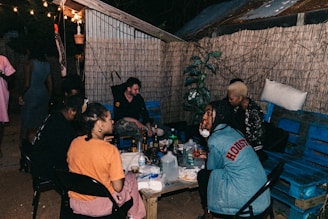 A small group of people sit around a low table outside, lit by string lights hanging above. They are in a casual setting with wooden walls and plants in the background. The table is covered with various drinks and snacks, and there are both men and women present, engaged in conversation and relaxed activities.