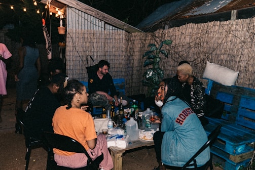 A small group of people sit around a low table outside, lit by string lights hanging above. They are in a casual setting with wooden walls and plants in the background. The table is covered with various drinks and snacks, and there are both men and women present, engaged in conversation and relaxed activities.