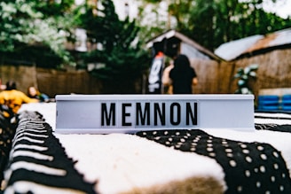 A focus on a lightbox displaying the word 'MEMNON' placed on a black and white patterned tablecloth, with an outdoor area and some people in the background, slightly out of focus, suggesting a casual gathering or event.