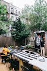 An outdoor dining area with a long table covered in patterned tablecloths, surrounded by black folding chairs. Two people are seated at the table, while another person is standing near a banner that reads 'Tangene House,' attached to a wooden structure. The backdrop includes trees, a wooden fence, and apartment buildings with fire escapes. There are various small potted plants and string lights above the area.