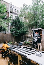 An outdoor dining area with a long table covered in patterned tablecloths, surrounded by black folding chairs. Two people are seated at the table, while another person is standing near a banner that reads 'Tangene House,' attached to a wooden structure. The backdrop includes trees, a wooden fence, and apartment buildings with fire escapes. There are various small potted plants and string lights above the area.
