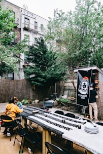 An outdoor dining area with a long table covered in patterned tablecloths, surrounded by black folding chairs. Two people are seated at the table, while another person is standing near a banner that reads 'Tangene House,' attached to a wooden structure. The backdrop includes trees, a wooden fence, and apartment buildings with fire escapes. There are various small potted plants and string lights above the area.