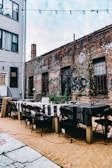 An outdoor setting with a long table and several black folding chairs arranged around it. The table is covered with patterned black and white tablecloths. The setting is in a narrow alley between two buildings. One of the buildings is made of brick and covered with graffiti, while the other appears to be a residential building with a gray exterior. String lights are hung above, and there are cables or wires lying on the ground.