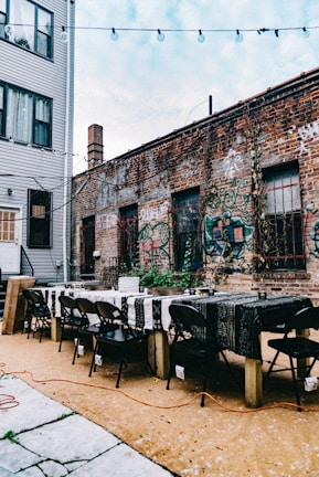 An outdoor setting with a long table and several black folding chairs arranged around it. The table is covered with patterned black and white tablecloths. The setting is in a narrow alley between two buildings. One of the buildings is made of brick and covered with graffiti, while the other appears to be a residential building with a gray exterior. String lights are hung above, and there are cables or wires lying on the ground.