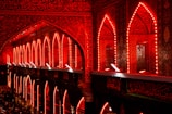 An architectural interior illuminated with vibrant red lighting, featuring intricately designed arches and walls covered in detailed patterns and Arabic calligraphy. Crowds of people can be seen gathered at the bottom, likely engaged in a religious or cultural event.