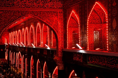 An architectural interior illuminated with vibrant red lighting, featuring intricately designed arches and walls covered in detailed patterns and Arabic calligraphy. Crowds of people can be seen gathered at the bottom, likely engaged in a religious or cultural event.