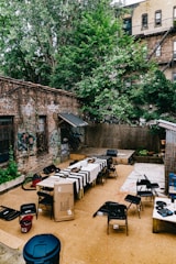 An outdoor urban setting with a fenced-in courtyard surrounded by brick buildings and a large tree. The courtyard is set up for a gathering, featuring several tables lined with black and white striped tablecloths and various serving trays. Black folding chairs are arranged around the tables, and there is a cardboard box nearby. The walls have graffiti, and there is visible electrical wiring extending to different parts of the area.