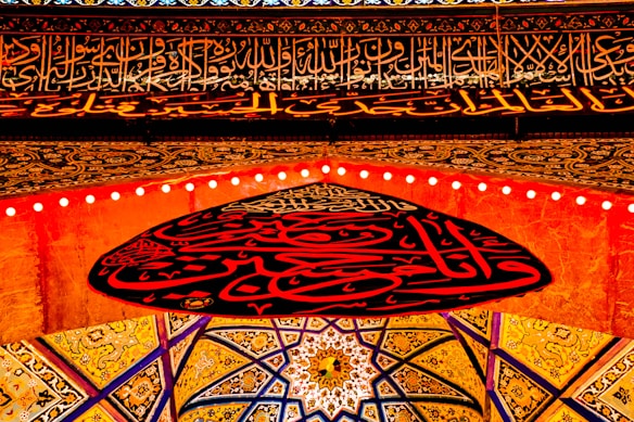 Ornate Islamic calligraphy and intricate geometric patterns decorate a ceiling. The design features vibrant red and gold colors, with Arabic script as a central element. The patterns create a symmetrical and harmonious visual effect.