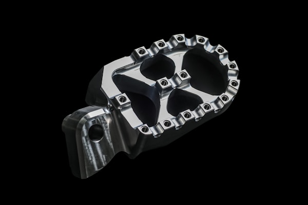 A close-up image of a metallic, machined part with a geometric design and bolts around the edges, placed against a black background.