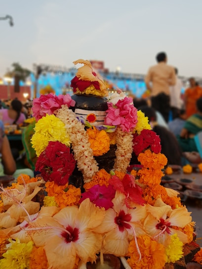 A beautifully decorated kalasha with a variety of colorful flowers including marigolds and hibiscus. The kalasha is adorned with garlands and placed amongst a crowd of people, suggesting a festive or religious gathering. The background reveals an outdoor setting with a blue sky and some blurry figures.