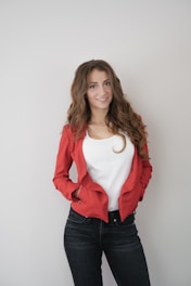 A woman with long, wavy brown hair wearing a red blazer over a white top and dark jeans. She is smiling and has her hands in her pockets against a plain light background.