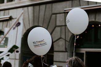 Two white balloons float above a crowd with a focus on their printed text that reads 'Wir Frauen* haben Recht!' The background showcases an urban setting with a building facade and a partially visible flag.
