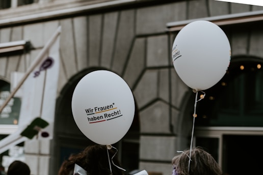Two white balloons float above a crowd with a focus on their printed text that reads 'Wir Frauen* haben Recht!' The background showcases an urban setting with a building facade and a partially visible flag.