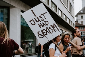 A group of people participating in a protest. A person holds a sign reading 'Just Had Sexism' while standing among other demonstrators. The setting appears to be an urban street with modern buildings in the background. The crowd is engaged and appears diverse in terms of gender.