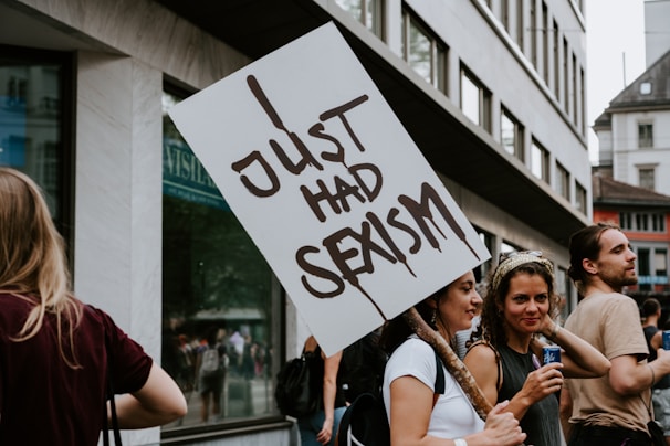 A group of people participating in a protest. A person holds a sign reading 'Just Had Sexism' while standing among other demonstrators. The setting appears to be an urban street with modern buildings in the background. The crowd is engaged and appears diverse in terms of gender.