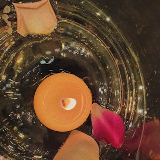 A lit tealight candle is placed in a shallow glass dish surrounded by delicate rose petals. The candle emits a warm, gentle glow, reflecting off the glossy surface beneath it, creating shimmering light patterns. The petals are in hues of pink and peach, adding a touch of elegance and romance to the scene.