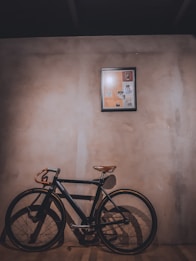 A vintage-style bicycle with a leather saddle and handlebars leans against a textured wall. Above it, a framed collage or artwork with various images is mounted. The lighting casts a warm shadow on the wooden floor, creating an intimate and nostalgic atmosphere.