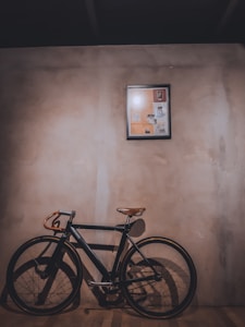 A vintage-style bicycle with a leather saddle and handlebars leans against a textured wall. Above it, a framed collage or artwork with various images is mounted. The lighting casts a warm shadow on the wooden floor, creating an intimate and nostalgic atmosphere.