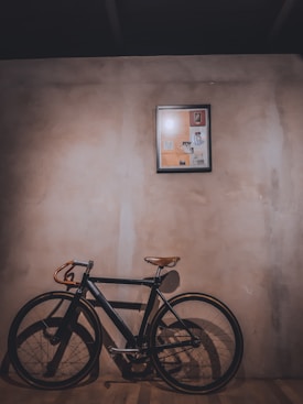 A vintage-style bicycle with a leather saddle and handlebars leans against a textured wall. Above it, a framed collage or artwork with various images is mounted. The lighting casts a warm shadow on the wooden floor, creating an intimate and nostalgic atmosphere.