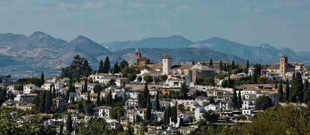 A picturesque town set against a backdrop of rolling hills and mountains, featuring clustered white buildings with terracotta roofs interspersed with tall cypress trees. The townscape includes churches with tall bell towers and a variety of vegetation around the structures.