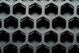 A close-up view of a hexagonal metal mesh structure with a textured, metallic surface. The hexagons are evenly distributed, creating a repetitive geometric pattern.