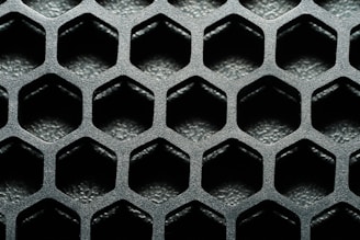 A close-up view of a hexagonal metal mesh structure with a textured, metallic surface. The hexagons are evenly distributed, creating a repetitive geometric pattern.