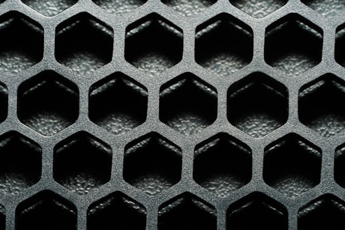A close-up view of a hexagonal metal mesh structure with a textured, metallic surface. The hexagons are evenly distributed, creating a repetitive geometric pattern.