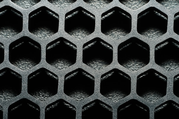 A close-up view of a hexagonal metal mesh structure with a textured, metallic surface. The hexagons are evenly distributed, creating a repetitive geometric pattern.