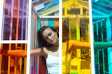 A woman stands with her arms outstretched inside a structure made of multicolored glass panels creating a vibrant and dynamic environment. Her expression is serene and slightly pensive as sunlight illuminates her face and the translucent colors cast intricate patterns of light and shadow.