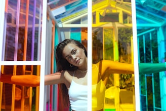 A woman stands with her arms outstretched inside a structure made of multicolored glass panels creating a vibrant and dynamic environment. Her expression is serene and slightly pensive as sunlight illuminates her face and the translucent colors cast intricate patterns of light and shadow.